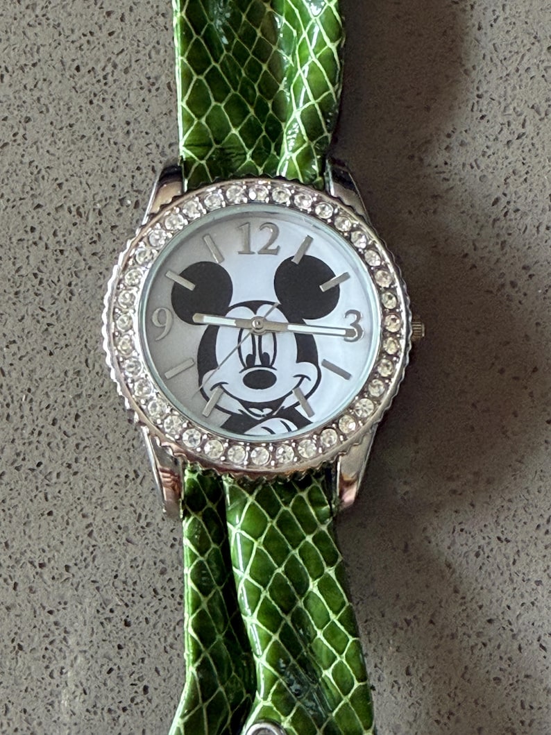 May include: A silver wristwatch with a white face featuring a Mickey Mouse design. The watch has a bezel encrusted with small, clear stones and a green, textured band. The watch face displays the numbers 12, 3, and 9.