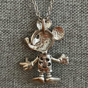 May include: Silver-coloured Mickey Mouse pendant necklace. The pendant features a detailed, three-dimensional design of Mickey Mouse with his arms outstretched. The necklace has a delicate chain and a lobster clasp.