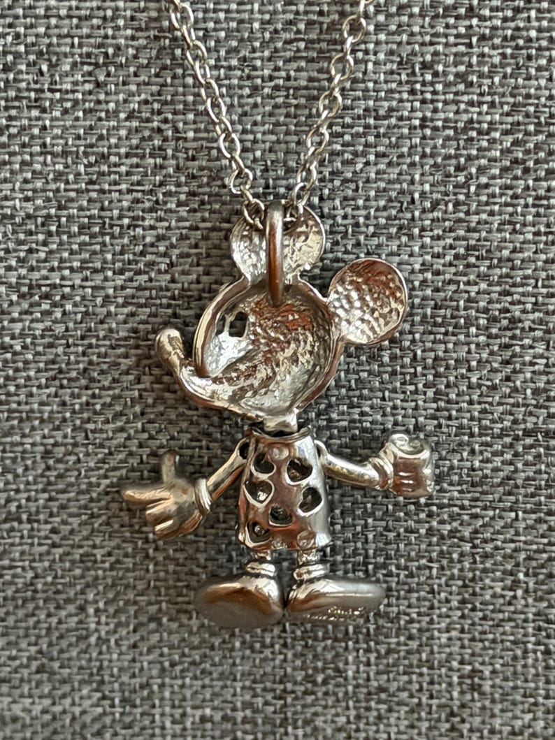 May include: Silver-coloured Mickey Mouse pendant necklace. The pendant features a detailed, three-dimensional design of Mickey Mouse with his arms outstretched. The necklace has a delicate chain and a lobster clasp.