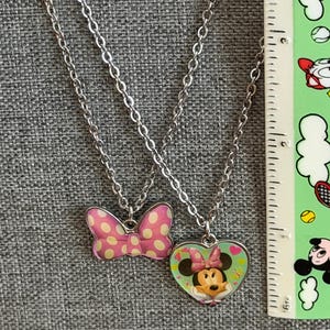 May include: Two silver-toned necklaces. One features a pink bow pendant with white polka dots. The other has a heart-shaped pendant with a Minnie Mouse graphic. The necklaces are displayed on a gray surface.