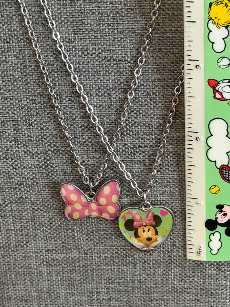 May include: Two silver-toned necklaces. One features a pink bow pendant with white polka dots. The other has a heart-shaped pendant with a Minnie Mouse graphic. The necklaces are displayed on a gray surface.