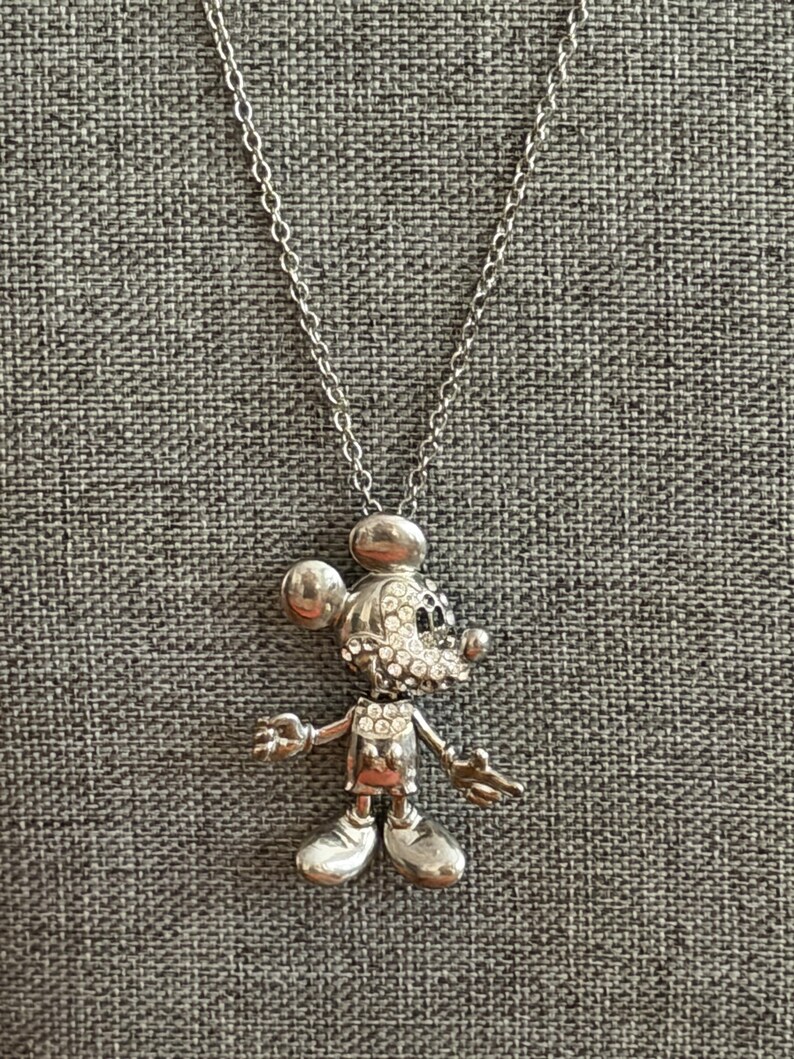 May include: Silver-coloured Mickey Mouse pendant necklace. The pendant features a detailed figure of Mickey Mouse, with sparkling crystal accents on the face. The necklace has a delicate chain and a lobster clasp.