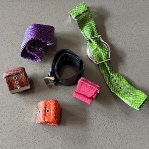 May include: A collection of colorful, textured leather accessories, including watch straps, bracelets, and a belt. The items are in shades of purple, brown, orange, pink, black, and green. The watch strap has a silver watch face.