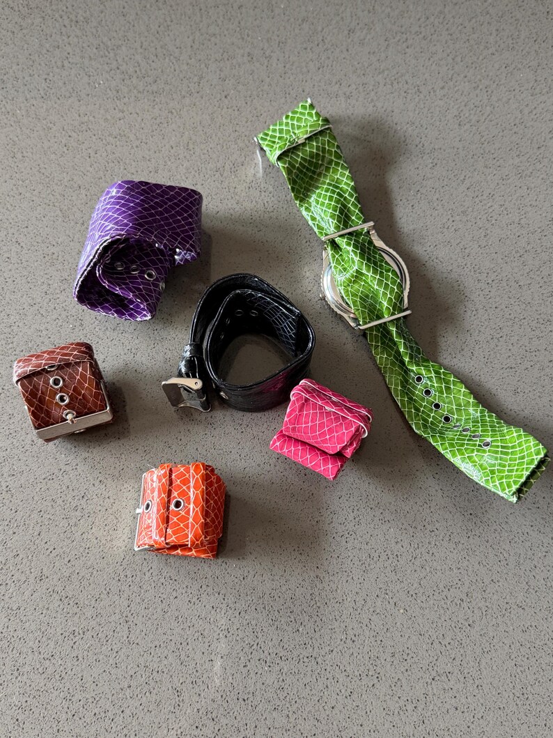 May include: A collection of colorful, textured leather accessories, including watch straps, bracelets, and a belt. The items are in shades of purple, brown, orange, pink, black, and green. The watch strap has a silver watch face.