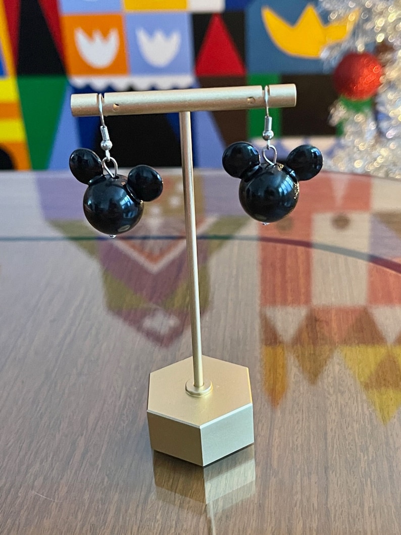 Puede incluir: Un par de pendientes negros con forma de Mickey Mouse con ganchos plateados. Los pendientes se muestran en un soporte dorado.
