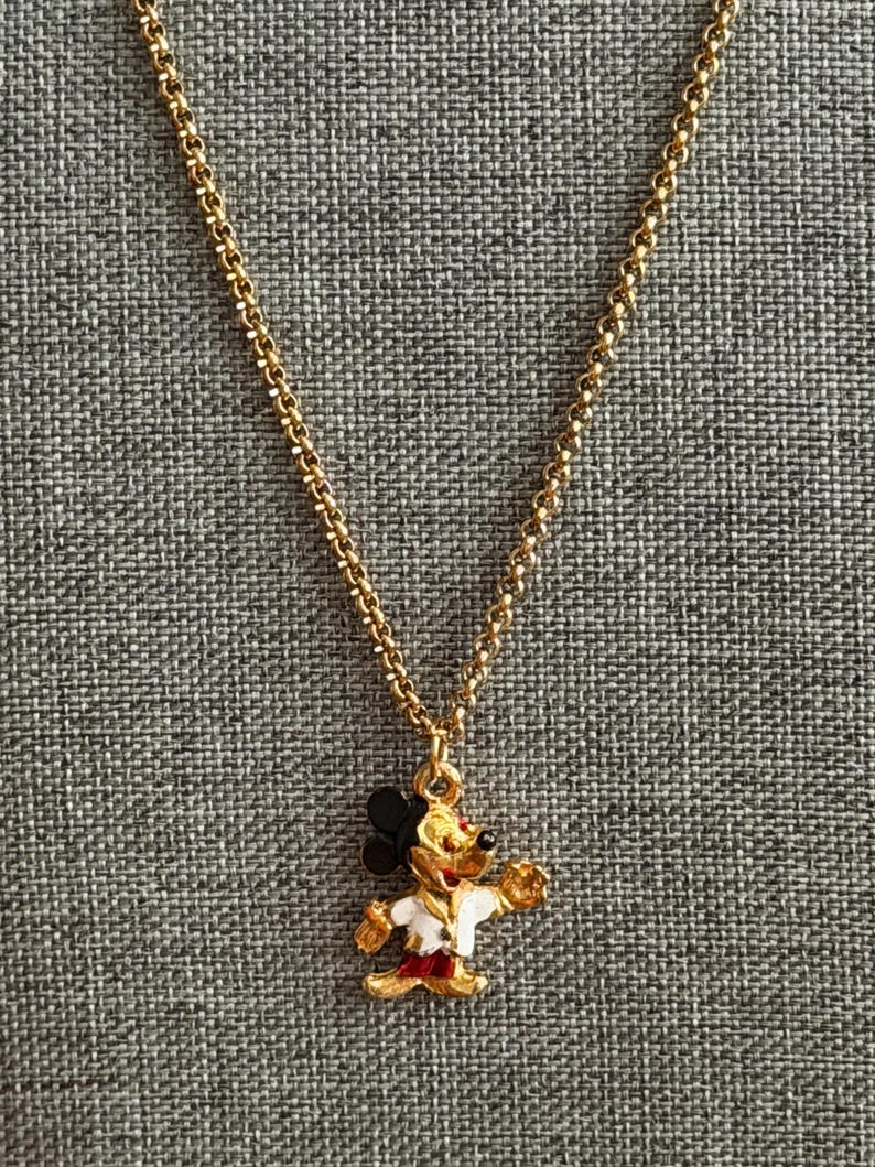 K&ouml;nnte beinhalten: Goldfarbene Halskette mit einem Mickey Mouse-Anh&auml;nger. Der Anh&auml;nger ist ein farbenfrohes Emaille-Design von Mickey Mouse in einer wei&szlig;en Jacke, roten Shorts und schwarzen Ohren. Die Kette ist im filigranen Gliederstil gehalten.