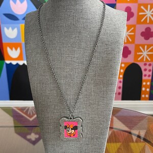 May include: A silver chain necklace with a silver frame pendant featuring a cartoon image of Minnie Mouse with a pink bow. The pendant is on a grey mannequin.