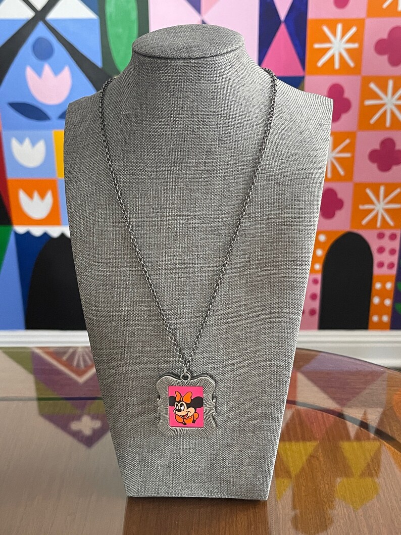 May include: A silver chain necklace with a silver frame pendant featuring a cartoon image of Minnie Mouse with a pink bow. The pendant is on a grey mannequin.