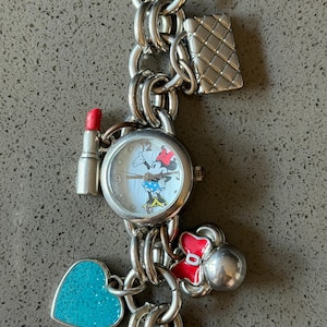 Puede incluir: Pulsera de dijes plateada con un reloj de Minnie Mouse, esfera blanca y lazo rojo. Los dijes incluyen un l&aacute;piz labial, un bolso acolchado, un coraz&oacute;n turquesa y un lazo rojo. La pulsera est&aacute; hecha de anillos de metal unidos.