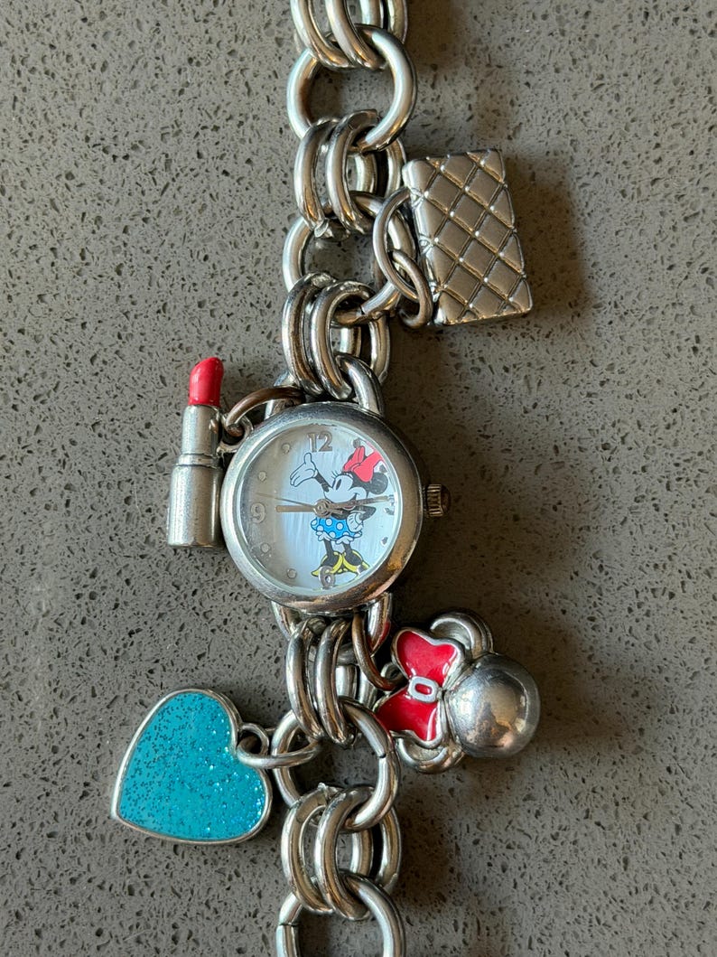 Puede incluir: Pulsera de dijes plateada con un reloj de Minnie Mouse, esfera blanca y lazo rojo. Los dijes incluyen un l&aacute;piz labial, un bolso acolchado, un coraz&oacute;n turquesa y un lazo rojo. La pulsera est&aacute; hecha de anillos de metal unidos.