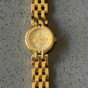 May include: Gold-coloured wristwatch with a rectangular link band. The watch face features a Mickey Mouse silhouette. The watch has a round face and a small crown. The watch band is made of gold-coloured metal.