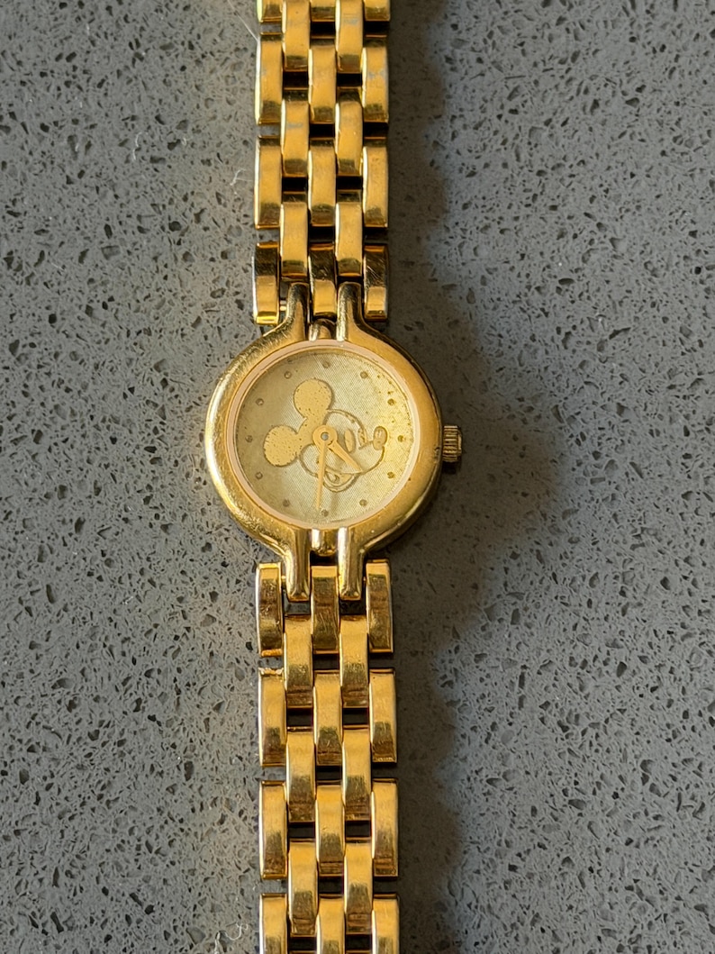 May include: Gold-coloured wristwatch with a rectangular link band. The watch face features a Mickey Mouse silhouette. The watch has a round face and a small crown. The watch band is made of gold-coloured metal.