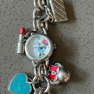 Puede incluir: Pulsera de dijes plateada con un reloj con dise&ntilde;o de Minnie Mouse, un dije de l&aacute;piz labial, un dije de bolso acolchado, un dije en forma de coraz&oacute;n y un dije de lazo rojo. La pulsera est&aacute; hecha de cadena enlazada.