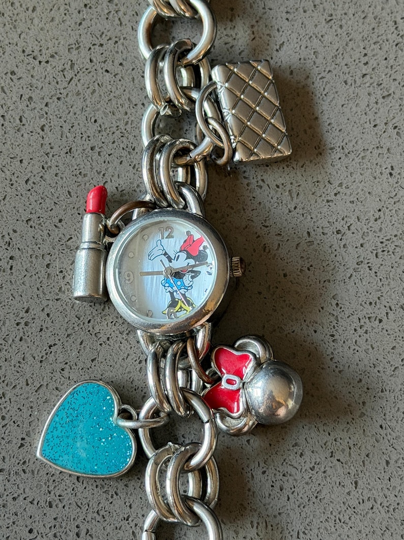 Puede incluir: Pulsera de dijes plateada con un reloj con dise&ntilde;o de Minnie Mouse, un dije de l&aacute;piz labial, un dije de bolso acolchado, un dije en forma de coraz&oacute;n y un dije de lazo rojo. La pulsera est&aacute; hecha de cadena enlazada.
