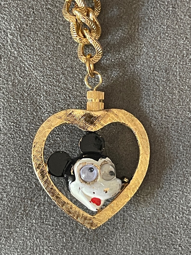 May include: A gold chain necklace with a heart-shaped pendant featuring a cartoon Mickey Mouse face with black ears, white face, and black eyes.