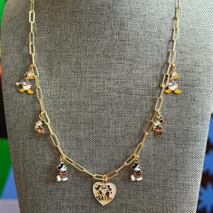 May include: Gold-tone chain necklace featuring Mickey Mouse, Minnie Mouse, and Donald Duck charms. The necklace includes a heart-shaped pendant with a Mickey and Minnie Mouse design. The charms are colorful and detailed, adding a playful touch to the jewelry.