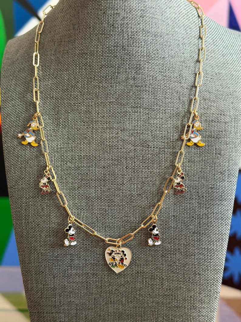 May include: Gold-tone chain necklace featuring Mickey Mouse, Minnie Mouse, and Donald Duck charms. The necklace includes a heart-shaped pendant with a Mickey and Minnie Mouse design. The charms are colorful and detailed, adding a playful touch to the jewelry.