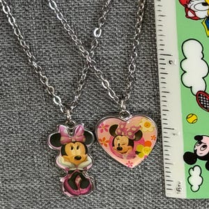 Peut inclure: Deux colliers argent&eacute;s avec des pendentifs Minnie Mouse. L'un est une figurine de Minnie Mouse, l'autre est un pendentif en forme de c&oelig;ur avec Minnie Mouse. Les pendentifs sont roses, blancs et noirs.