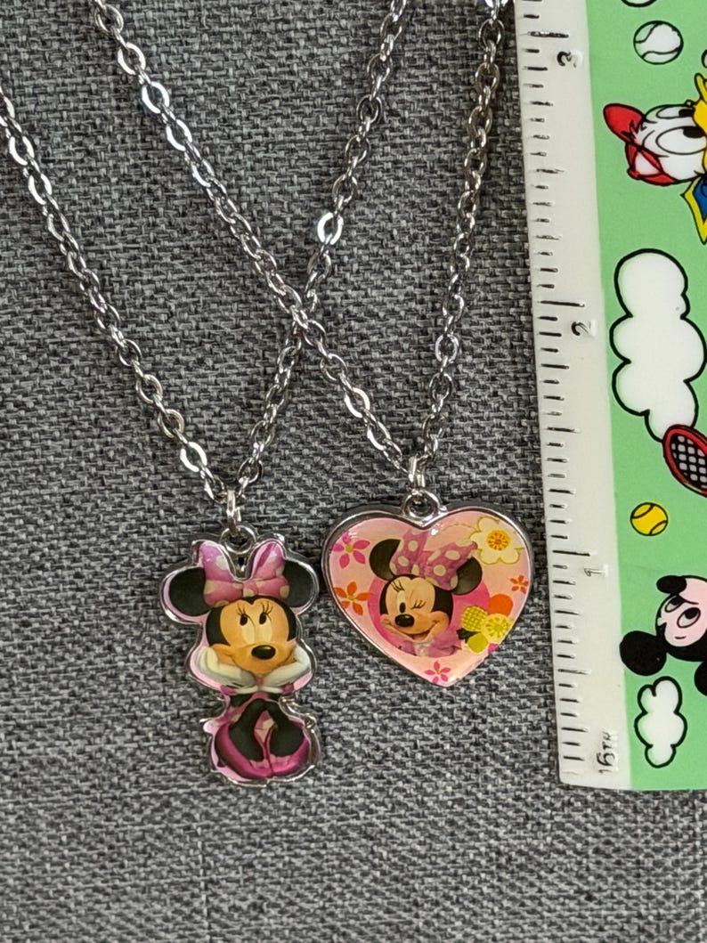 Peut inclure: Deux colliers argent&eacute;s avec des pendentifs Minnie Mouse. L'un est une figurine de Minnie Mouse, l'autre est un pendentif en forme de c&oelig;ur avec Minnie Mouse. Les pendentifs sont roses, blancs et noirs.