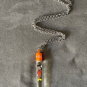 May include: A silver chain necklace with a clear plastic tube pendant. The tube features a cartoon image of Mickey Mouse in a red shirt and yellow shoes. The tube is topped with an orange cap that reads "Walt Prod."