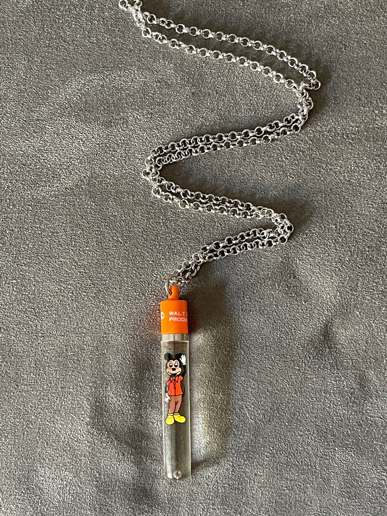 May include: A silver chain necklace with a clear plastic tube pendant. The tube features a cartoon image of Mickey Mouse in a red shirt and yellow shoes. The tube is topped with an orange cap that reads "Walt Prod."