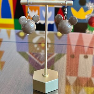 Puede incluir: Un par de pendientes marrones en forma de Mickey Mouse con ganchos plateados. Los pendientes se muestran en un soporte dorado sobre un fondo colorido.