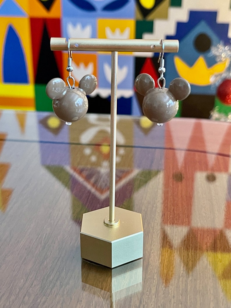Puede incluir: Un par de pendientes marrones en forma de Mickey Mouse con ganchos plateados. Los pendientes se muestran en un soporte dorado sobre un fondo colorido.
