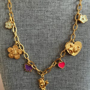 May include: Gold-toned necklace with multiple charms. Charms include flowers, a bow, a heart, and a character figure. The necklace has a chain link design and is displayed against a gray background.