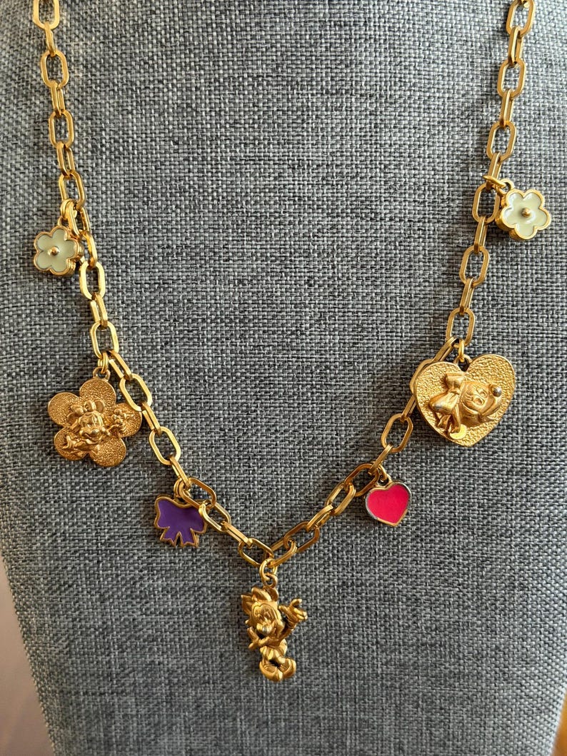 May include: Gold-toned necklace with multiple charms. Charms include flowers, a bow, a heart, and a character figure. The necklace has a chain link design and is displayed against a gray background.