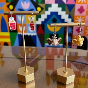 May include: Two pairs of earrings on a gold display stand. The first pair features a red and white popcorn bucket. The second pair features a brown and white popcorn bucket.