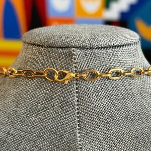 May include: A gold-colored necklace with a rectangular link chain and a lobster clasp closure. The necklace is displayed on a gray mannequin bust. The background is a colorful abstract design.