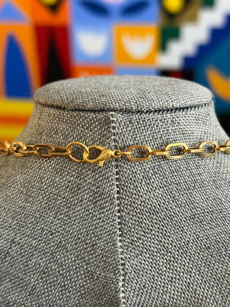 May include: A gold-colored necklace with a rectangular link chain and a lobster clasp closure. The necklace is displayed on a gray mannequin bust. The background is a colorful abstract design.