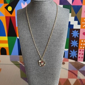 May include: A gold-colored necklace with a pendant featuring a Minnie Mouse design. The pendant has red, white, and black enamel details. The necklace is displayed on a gray mannequin head against a colorful background.