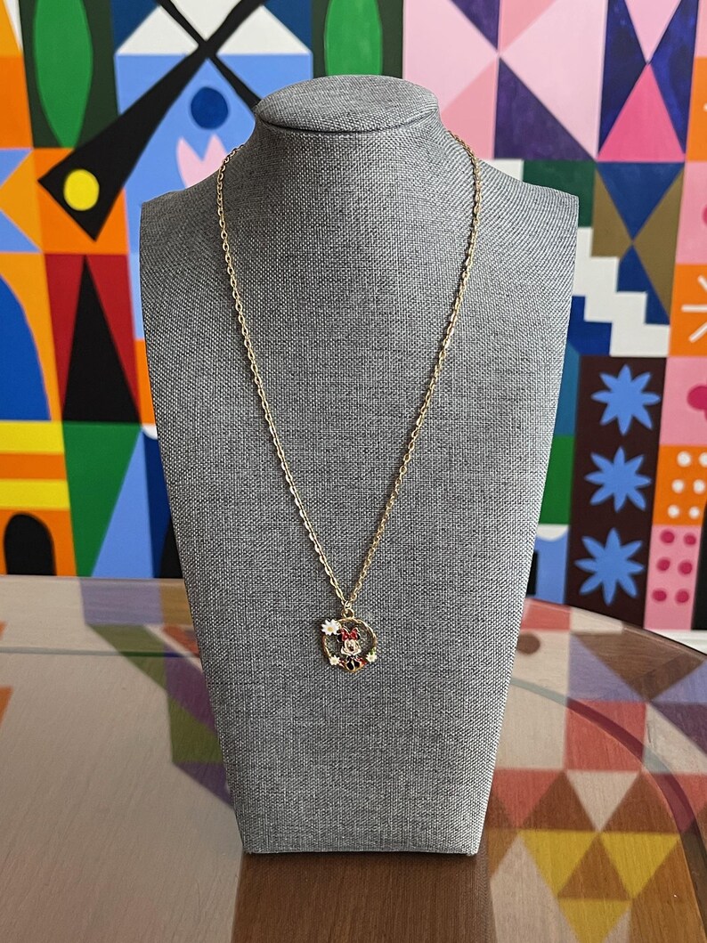 May include: A gold-colored necklace with a pendant featuring a Minnie Mouse design. The pendant has red, white, and black enamel details. The necklace is displayed on a gray mannequin head against a colorful background.
