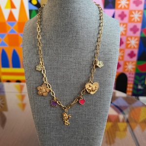 May include: Gold-toned necklace with multiple charms, including flowers, hearts, and a small figure. The necklace is displayed on a gray mannequin, showcasing its design and details. The charms are in various colors.