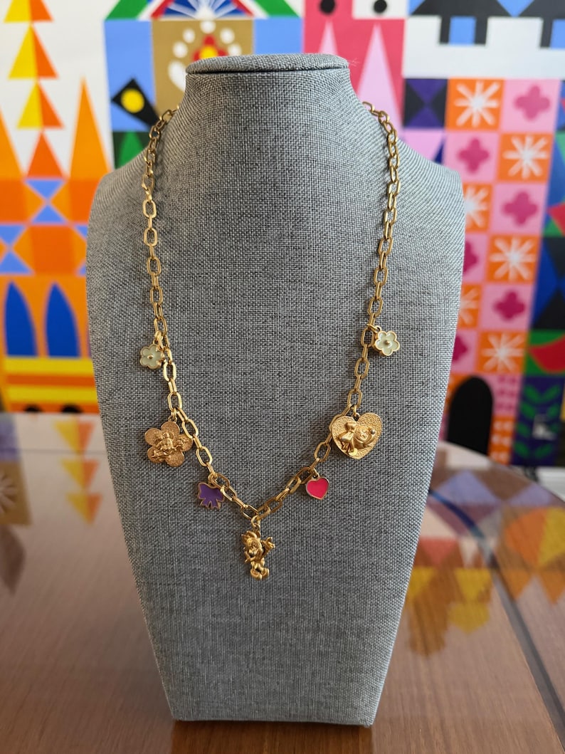 May include: Gold-toned necklace with multiple charms, including flowers, hearts, and a small figure. The necklace is displayed on a gray mannequin, showcasing its design and details. The charms are in various colors.