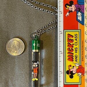 May include: A silver chain necklace with a green and clear glass pendant featuring a cartoon image of Mickey Mouse. The pendant is approximately 2 inches long. The text "WALT DISNEY PRODUCTIONS" is printed on the ruler in the background.