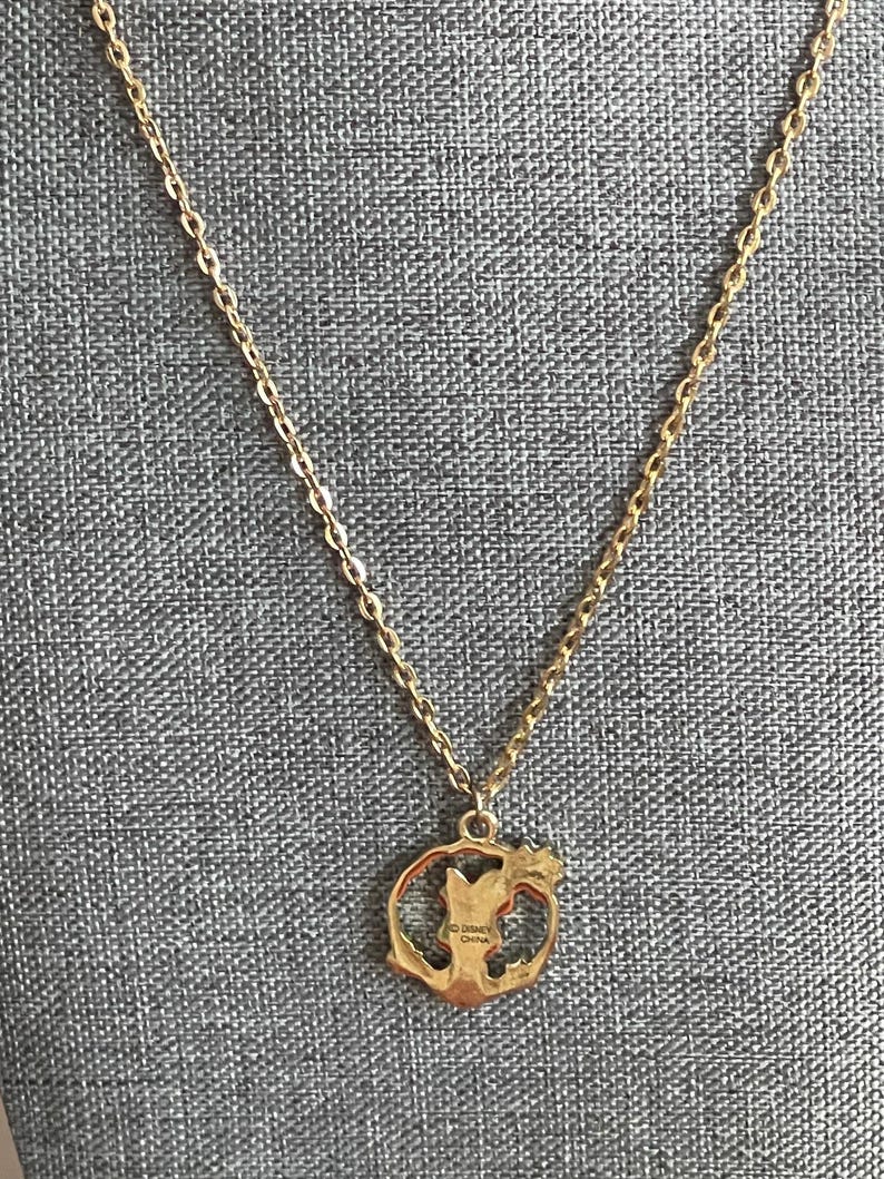 May include: Gold-toned necklace with a pendant featuring a character silhouette within a circular frame. The chain is delicate, and the pendant has the text "Disney China". The necklace is displayed against a textured gray background.