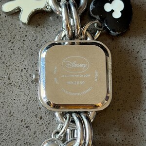 May include: A silver-toned Disney watch with a square face and a stainless steel back. The watch band features Mickey Mouse-themed charms, including a white glove and a black heart with white Mickey Mouse ears. The watch face reads "Disney" and "MK2059".