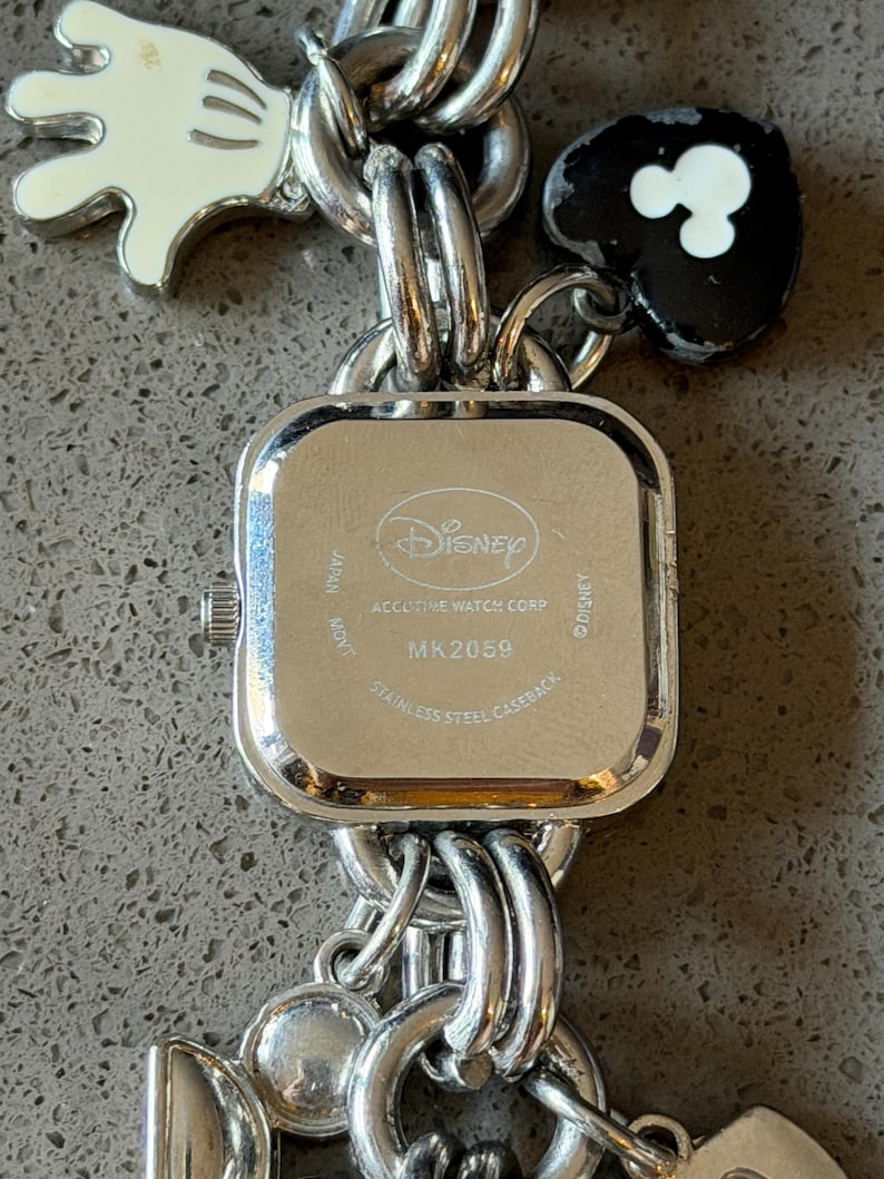 May include: A silver-toned Disney watch with a square face and a stainless steel back. The watch band features Mickey Mouse-themed charms, including a white glove and a black heart with white Mickey Mouse ears. The watch face reads "Disney" and "MK2059".