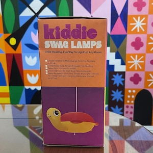 May include: A box for a Kiddie Swag Lamps turtle nightlight. The box is orange, purple, and yellow. The turtle is yellow and red. The box says "Kiddie Swag Lamps" and "Child Pleasing, Fun Way To Light Up Any Room".