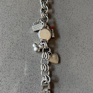 Puede incluir: Reloj de pulsera con abalorios plateado con esfera redonda y correa de cadena. Los abalorios incluyen un coraz&oacute;n, un l&aacute;piz labial, un bolso y abalorios con forma de Mickey Mouse. El reloj se muestra sobre una superficie gris.