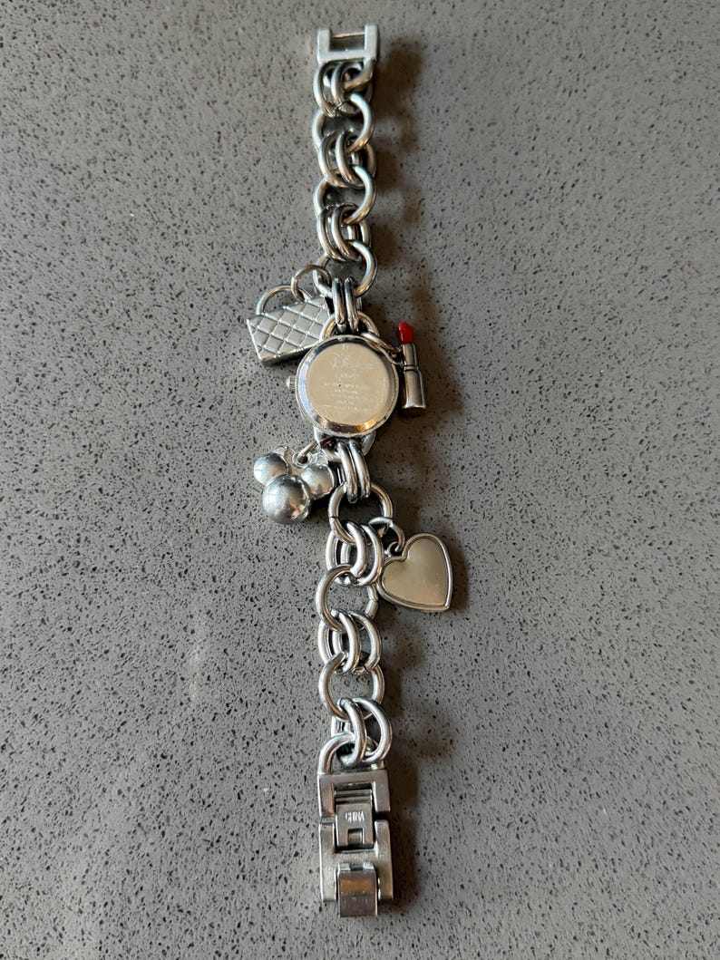 Puede incluir: Reloj de pulsera con abalorios plateado con esfera redonda y correa de cadena. Los abalorios incluyen un coraz&oacute;n, un l&aacute;piz labial, un bolso y abalorios con forma de Mickey Mouse. El reloj se muestra sobre una superficie gris.