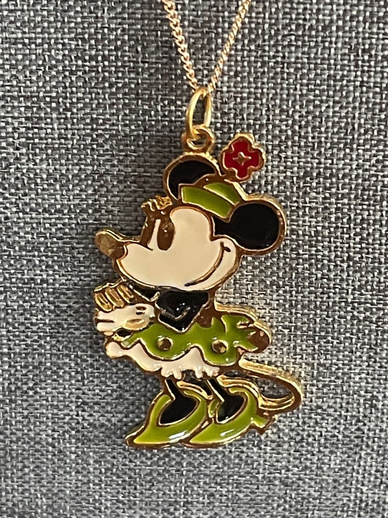 May include: A gold chain necklace with a green and black enamel Minnie Mouse pendant. The pendant features a red flower on her head and a white face.
