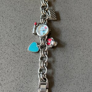Puede incluir: Reloj de pulsera con abalorios plateado con una esfera redonda con un personaje de dibujos animados. La pulsera tiene varios abalorios, como un l&aacute;piz labial, un coraz&oacute;n y un bolso. El reloj est&aacute; sobre una superficie gris.