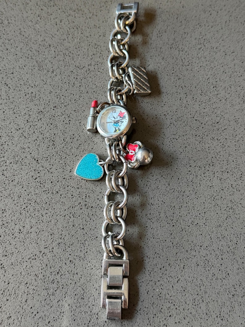 Puede incluir: Reloj de pulsera con abalorios plateado con una esfera redonda con un personaje de dibujos animados. La pulsera tiene varios abalorios, como un l&aacute;piz labial, un coraz&oacute;n y un bolso. El reloj est&aacute; sobre una superficie gris.