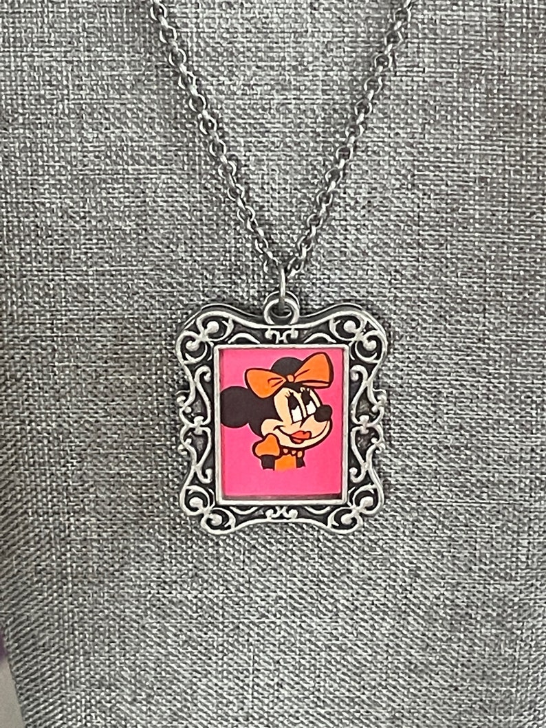 May include: A silver chain necklace with a silver picture frame pendant featuring a cartoon image of Minnie Mouse with a pink background.