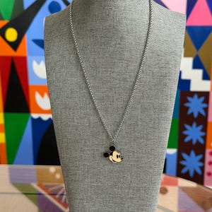 Pu&ograve; includere: Una collana color argento con un ciondolo di Topolino. Il ciondolo presenta una testa e orecchie nere, una bocca rossa e un viso giallo. La collana &egrave; esposta su un manichino grigio.