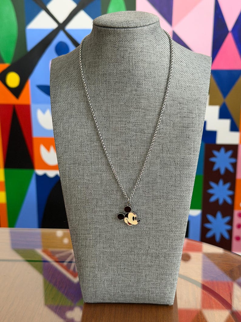 Pu&ograve; includere: Una collana color argento con un ciondolo di Topolino. Il ciondolo presenta una testa e orecchie nere, una bocca rossa e un viso giallo. La collana &egrave; esposta su un manichino grigio.