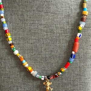 May include: A colorful beaded necklace with a gold-colored Mickey Mouse charm. The necklace features a variety of small, multi-colored beads, including red, yellow, blue, and white. The charm is a detailed figure of Mickey Mouse.